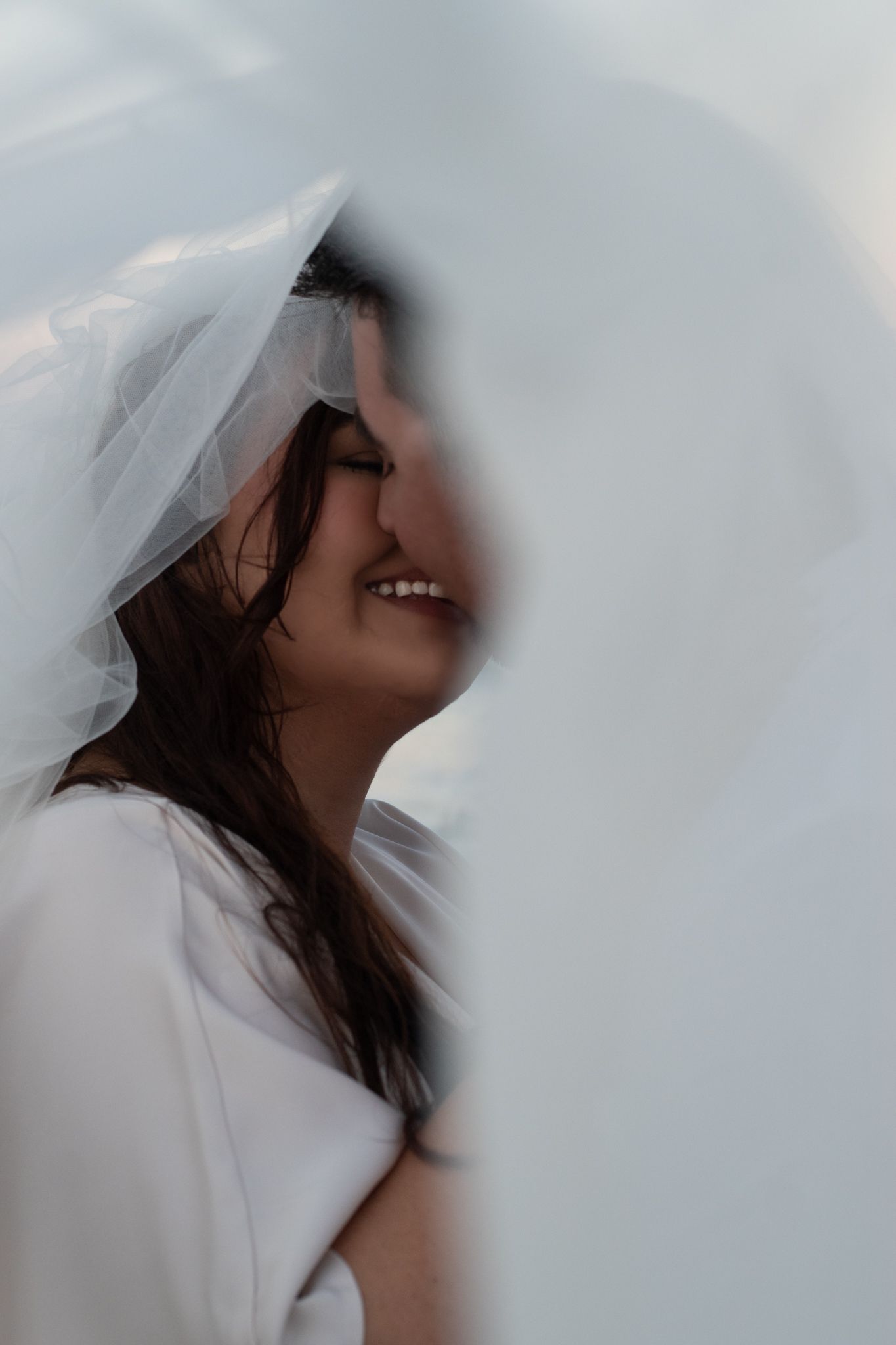 Side profile of person wearing white wedding veil smiling against light background.