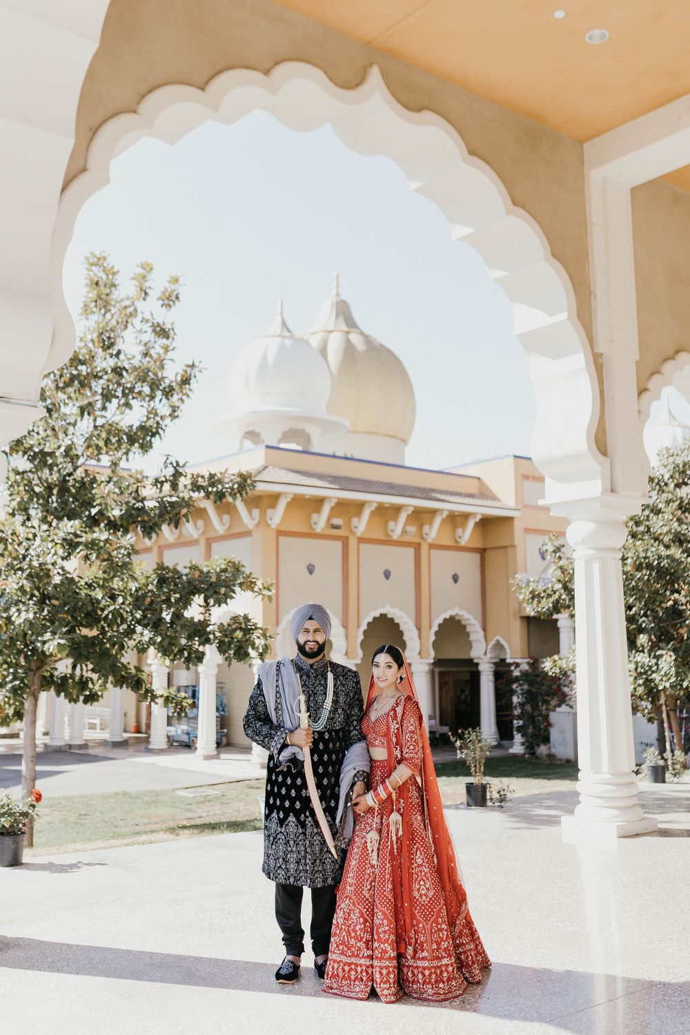 Indian wedding couple in traditional red and black attire standing under ornate white archway.