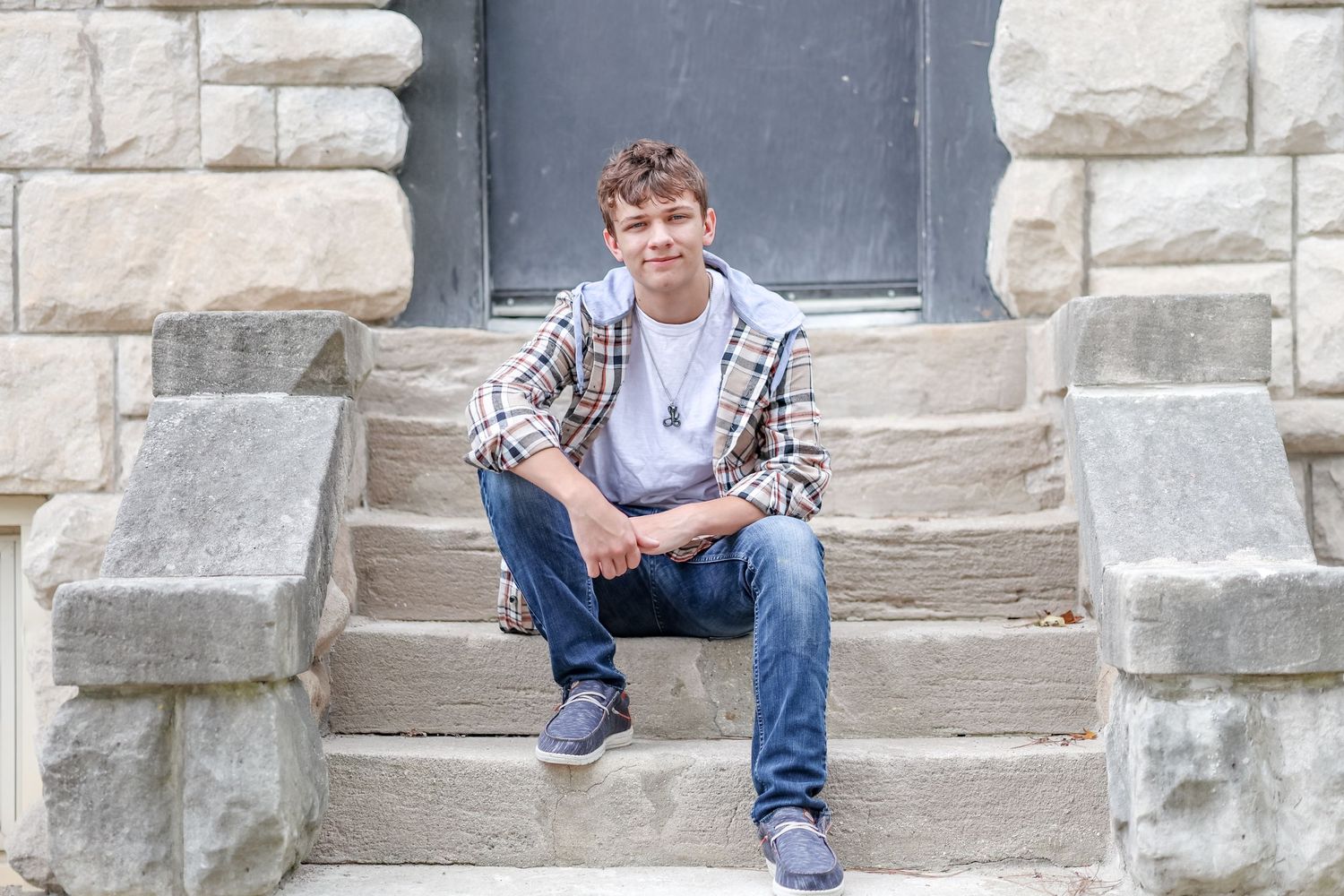 Senior in white shirt and tan plaid jacket leisurely sits on steps outside in front of a black door