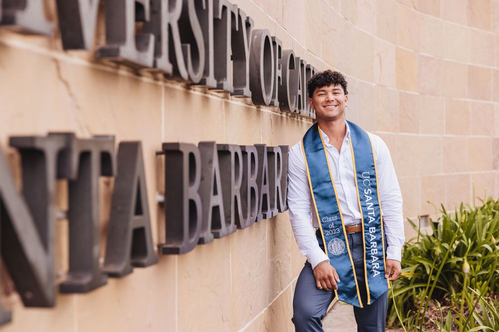 Graduate wearing white shirt and blue stole smiles while standing by campus wall.