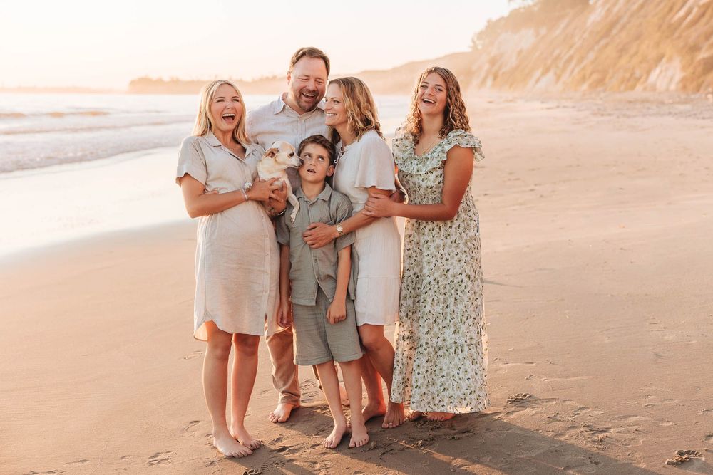 Family of five smiling and laughing together on a sandy beach at golden hour sunset.