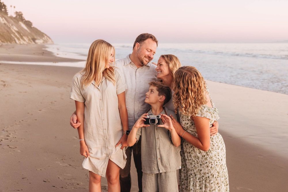 Family of five smiling together on a sandy beach at sunset, with a child holding a vintage camera.