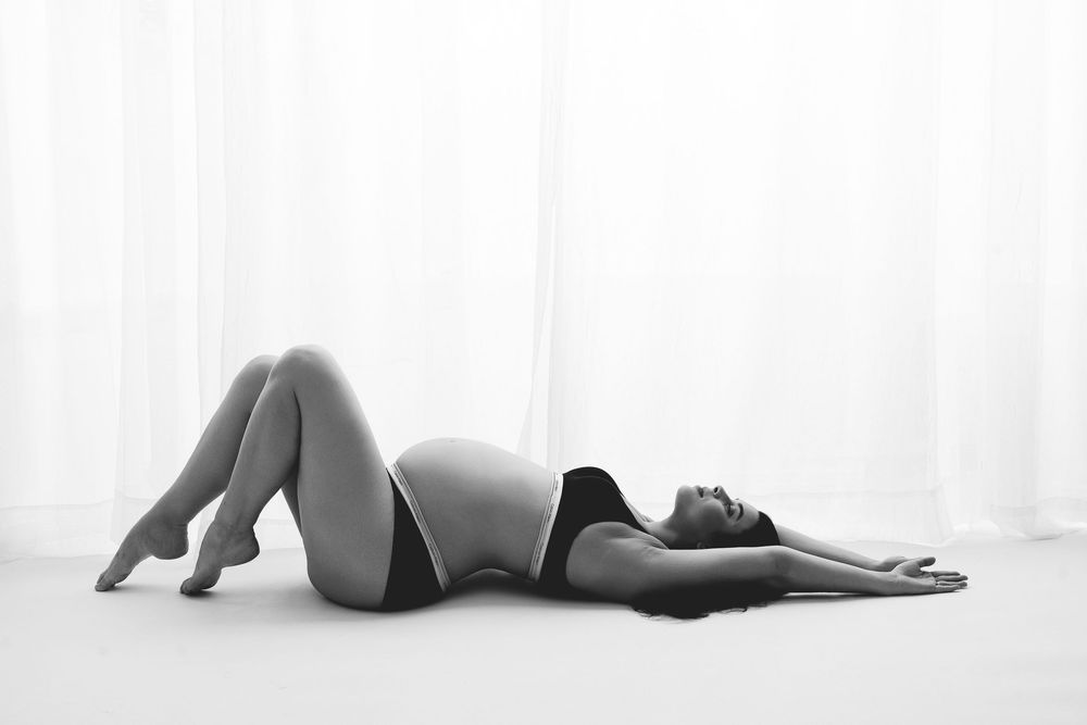 Black and white maternity photo of a pregnant woman lying on the floor in lingerie, posing gracefully.
