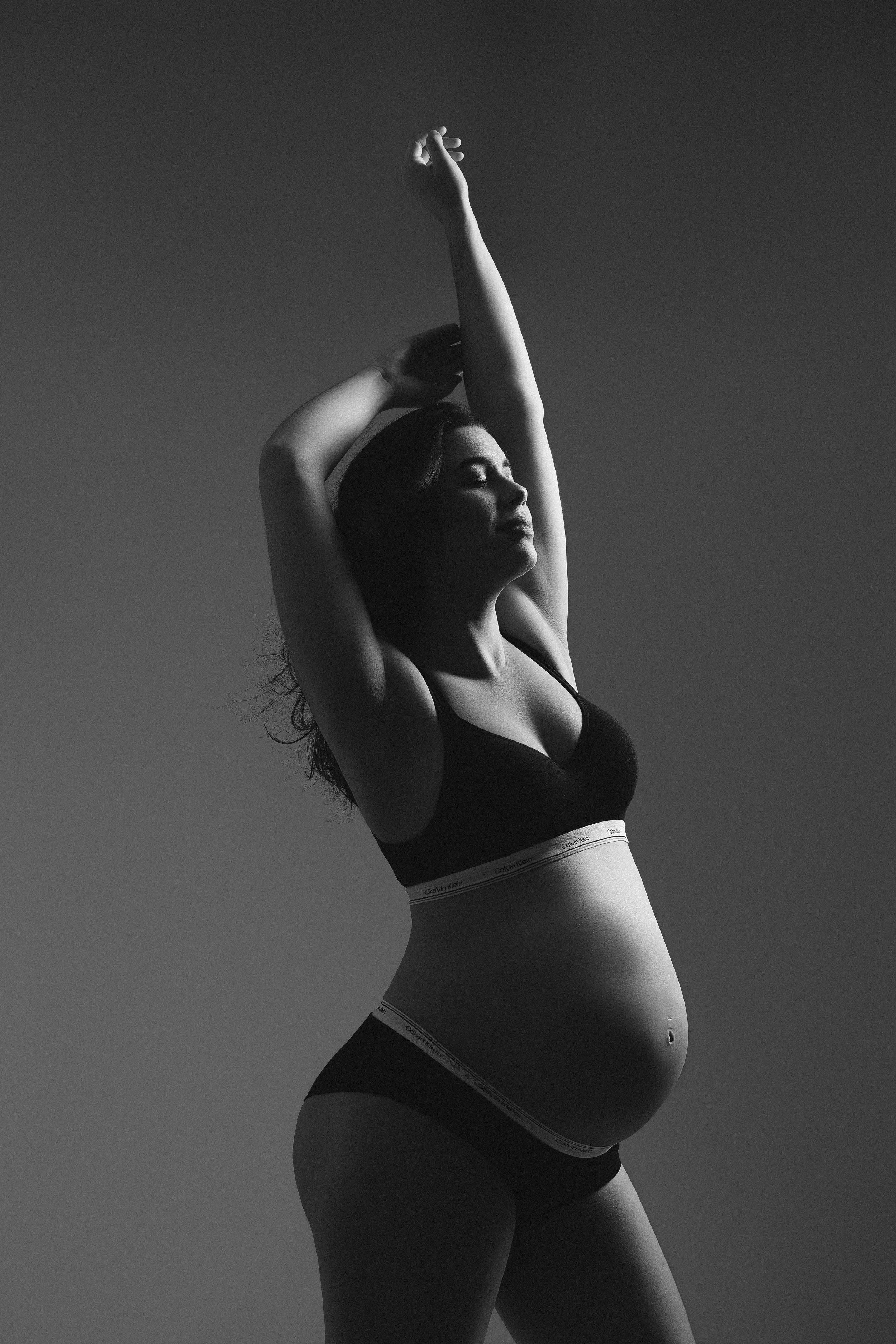 Black and white photo of a pregnant woman in lingerie stretching arms overhead, showcasing her baby bump.