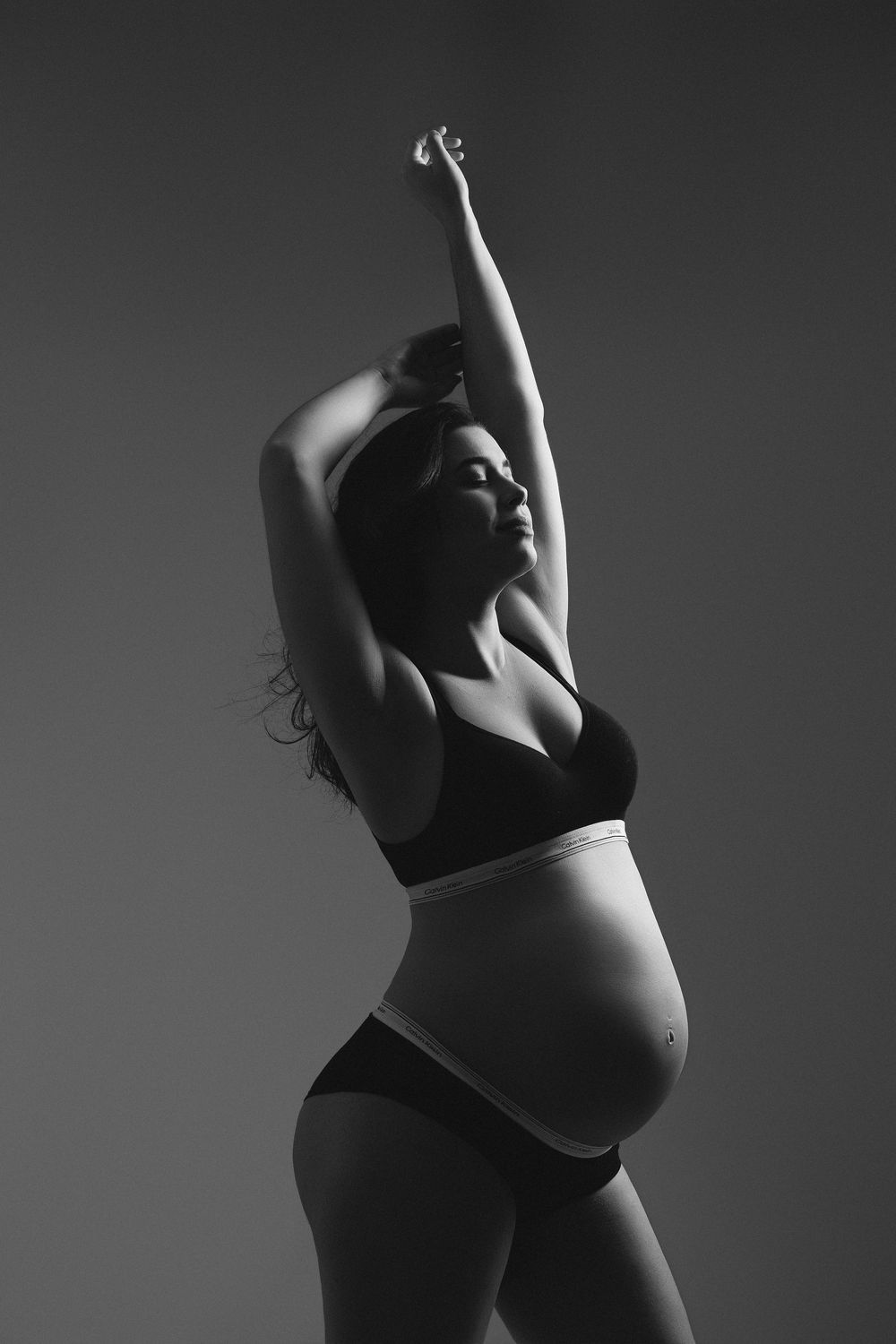 Black and white photo of a pregnant woman in lingerie stretching arms overhead, showcasing her baby bump.
