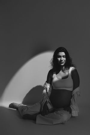 Black and white portrait of a woman with curly hair sitting cross-legged on floor in dramatic spotlight lighting.