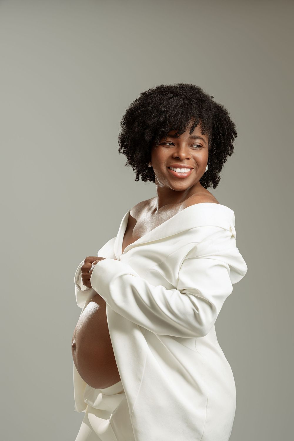 Smiling pregnant Black woman in white blazer showing baby bump, studio portrait with gray background.