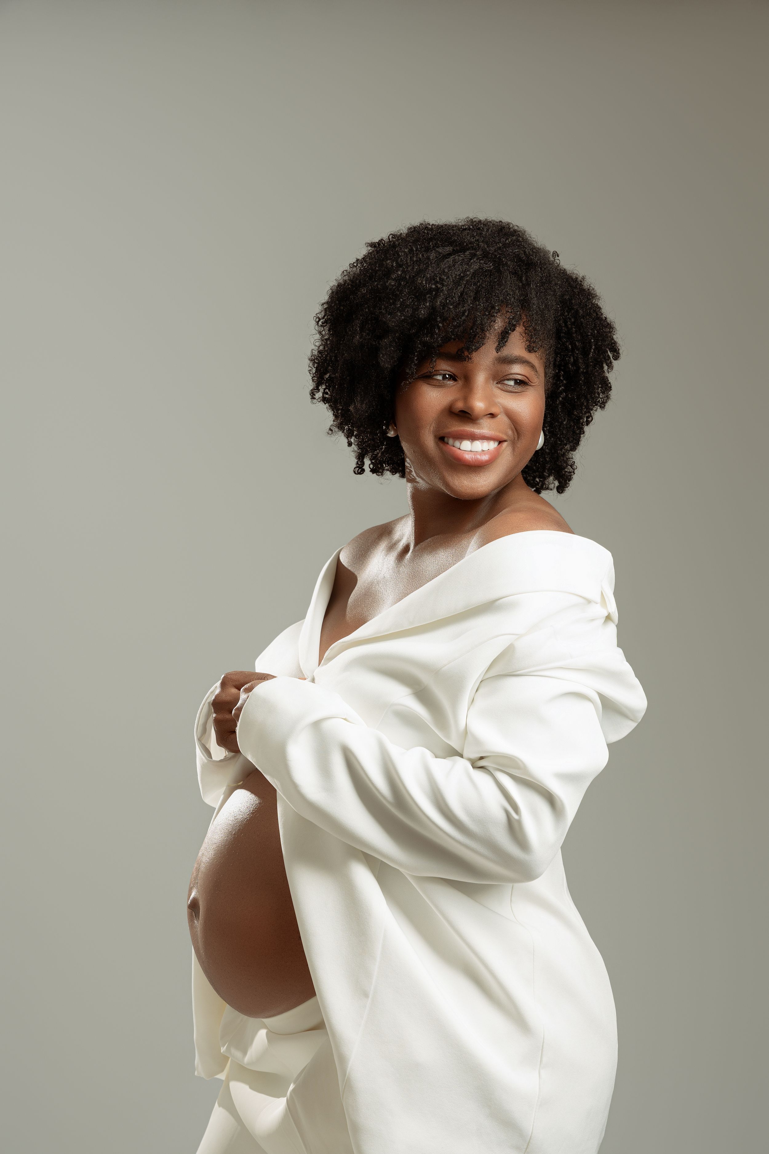 Smiling pregnant Black woman in white blazer showing baby bump, studio portrait with gray background.
