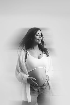 Black and white maternity photo of an expectant mother in a white crop top and jeans smiling with wind-blown hair. Paloma Schell