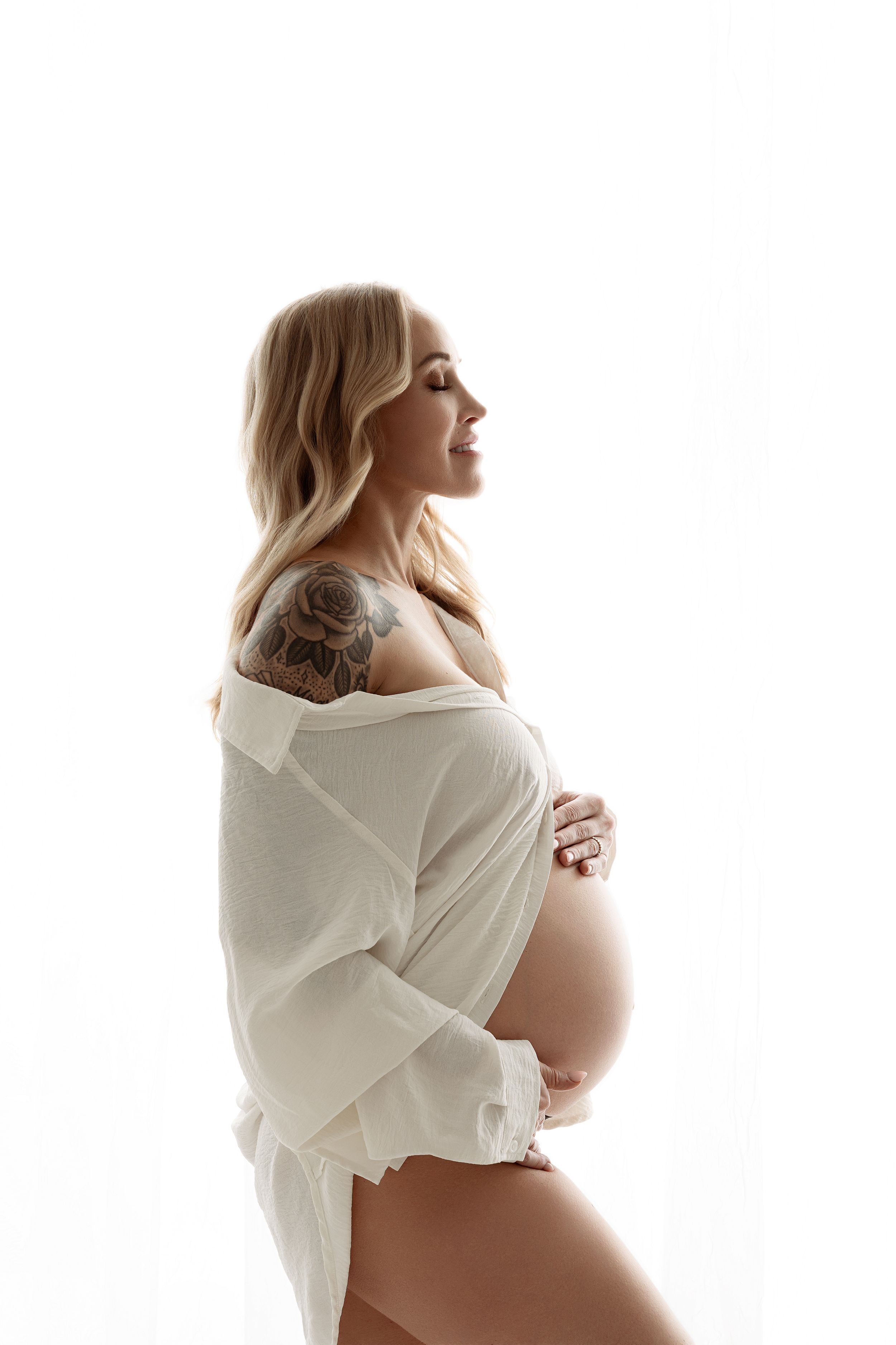 Pregnant woman in white open shirt cradling baby bump, showing shoulder tattoo, profile view on white background.