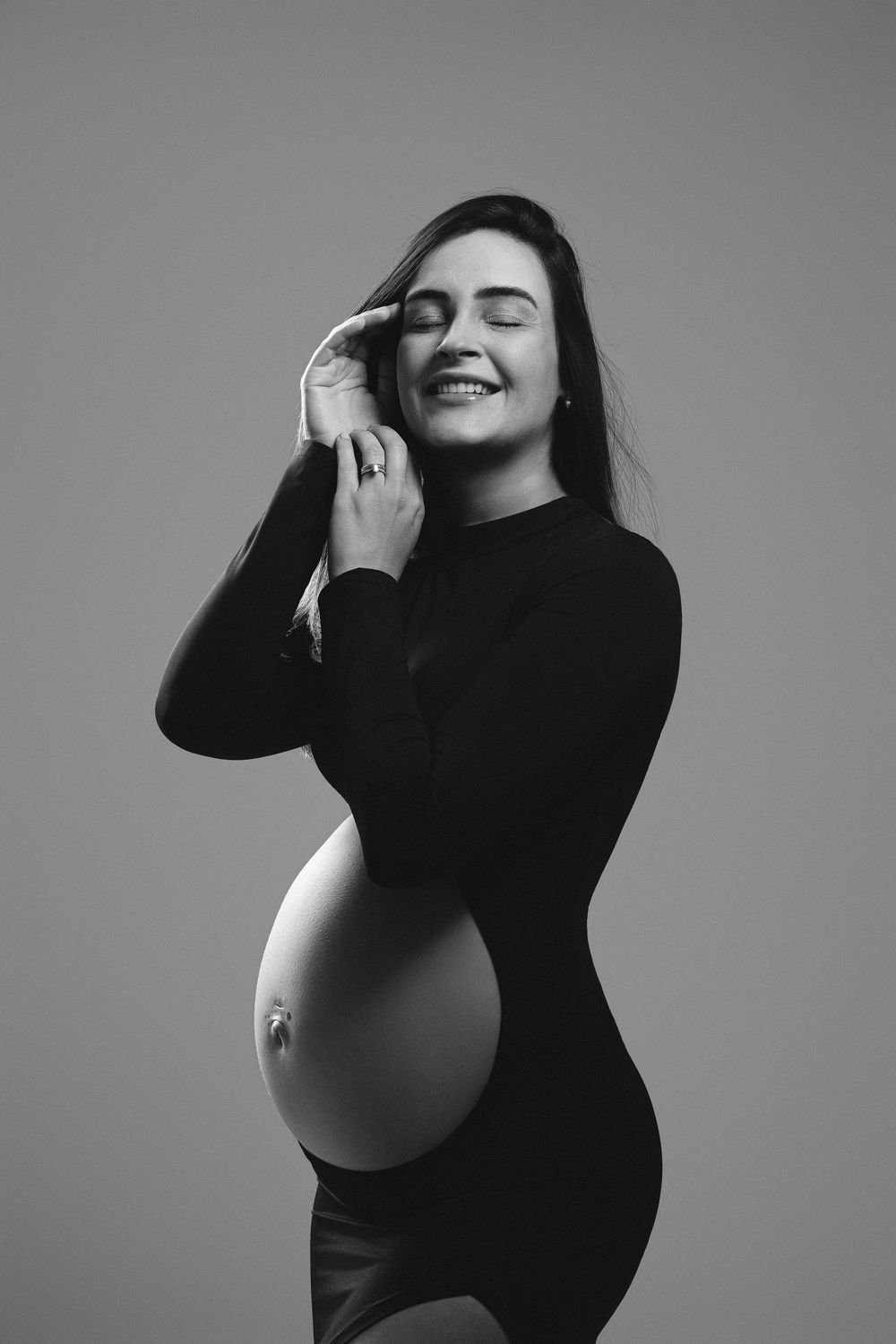 Black and white portrait of a smiling pregnant woman in a black dress, touching her face with eyes closed.