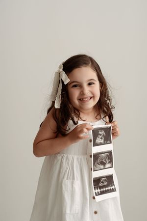 Smiling little girl in white dress holding ultrasound photos, announcing a new sibling.