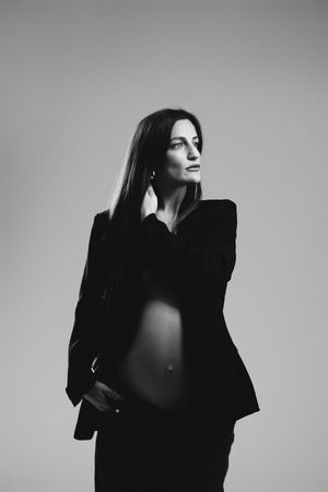 Black and white portrait of a pregnant woman in an open blazer, posing gracefully against a neutral background.