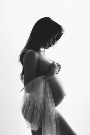 Black and white silhouette of a pregnant woman cradling her baby bump, draped in sheer fabric against white background.