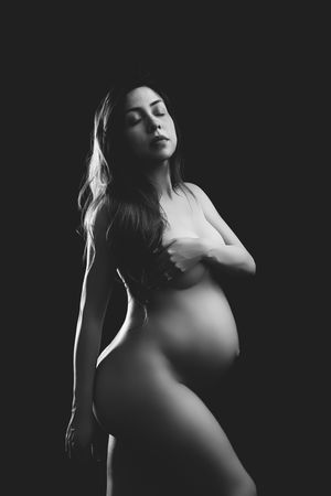 Black and white artistic maternity photo of pregnant woman posing gracefully against dark background.