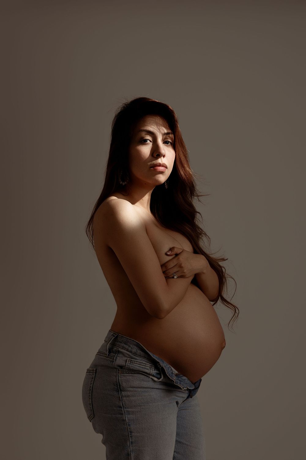 Pregnant woman with long hair posing topless in jeans, cradling her baby bump in a studio setting.