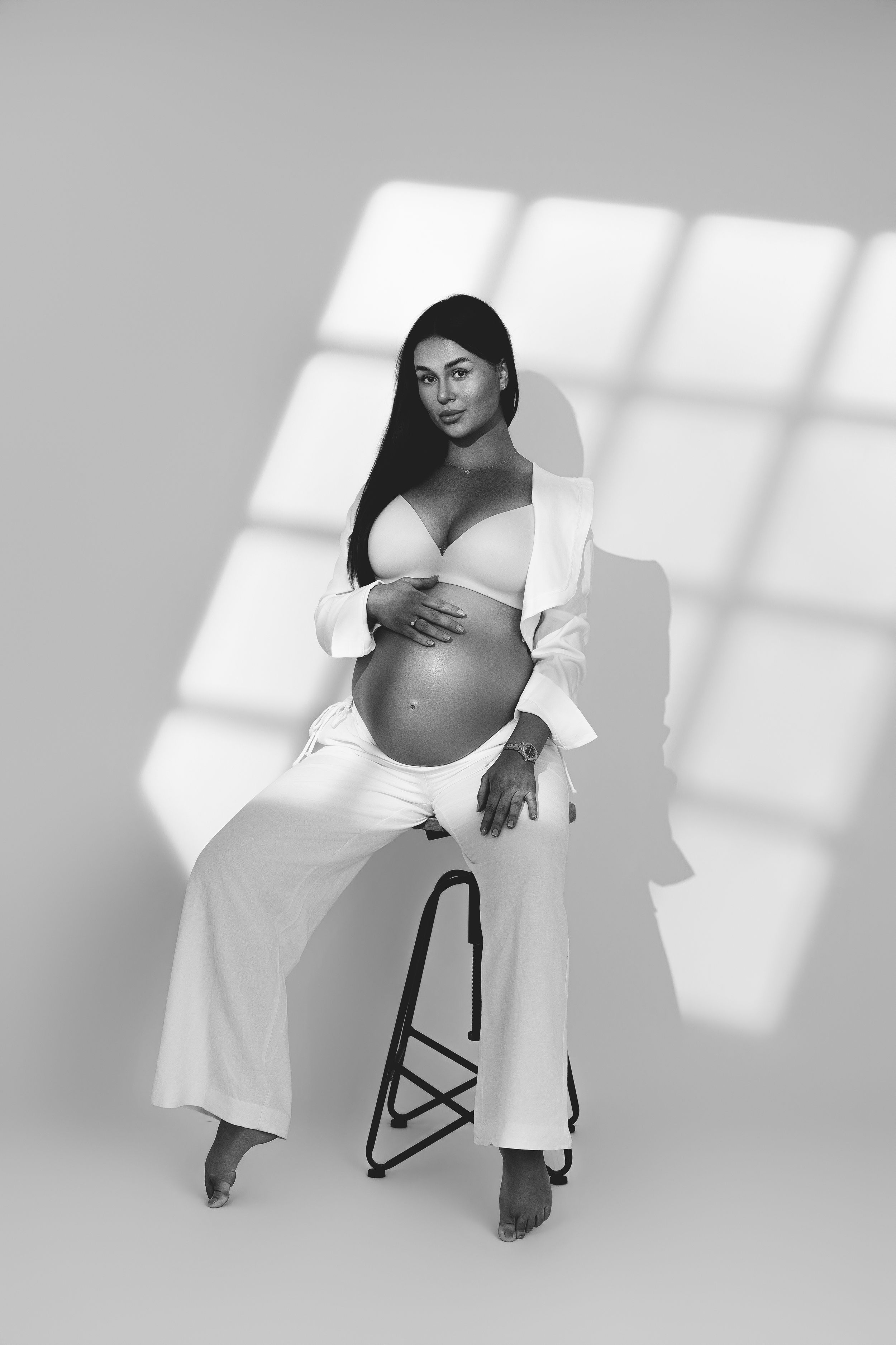 Black and white maternity photo of a pregnant woman in white outfit sitting on a stool, cradling her baby bump.