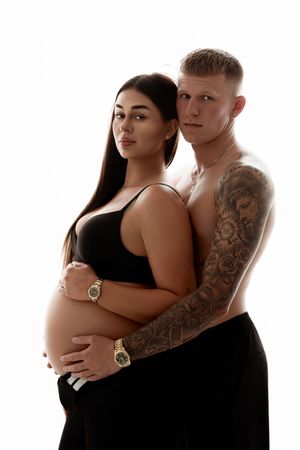 Pregnant couple posing together, shirtless tattooed man embracing woman with baby bump in black outfit.