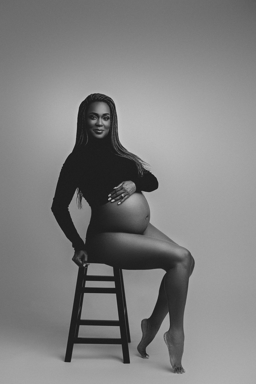 A black and white maternity photo with a person posing elegantly on a wooden stool.