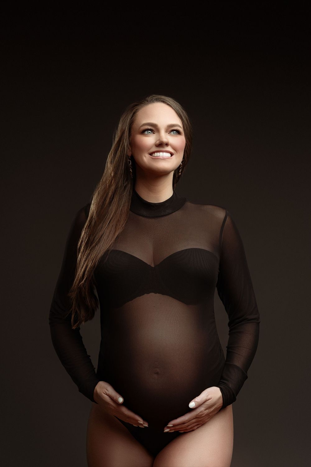 Maternity portrait in black mesh bodysuit against dark background.