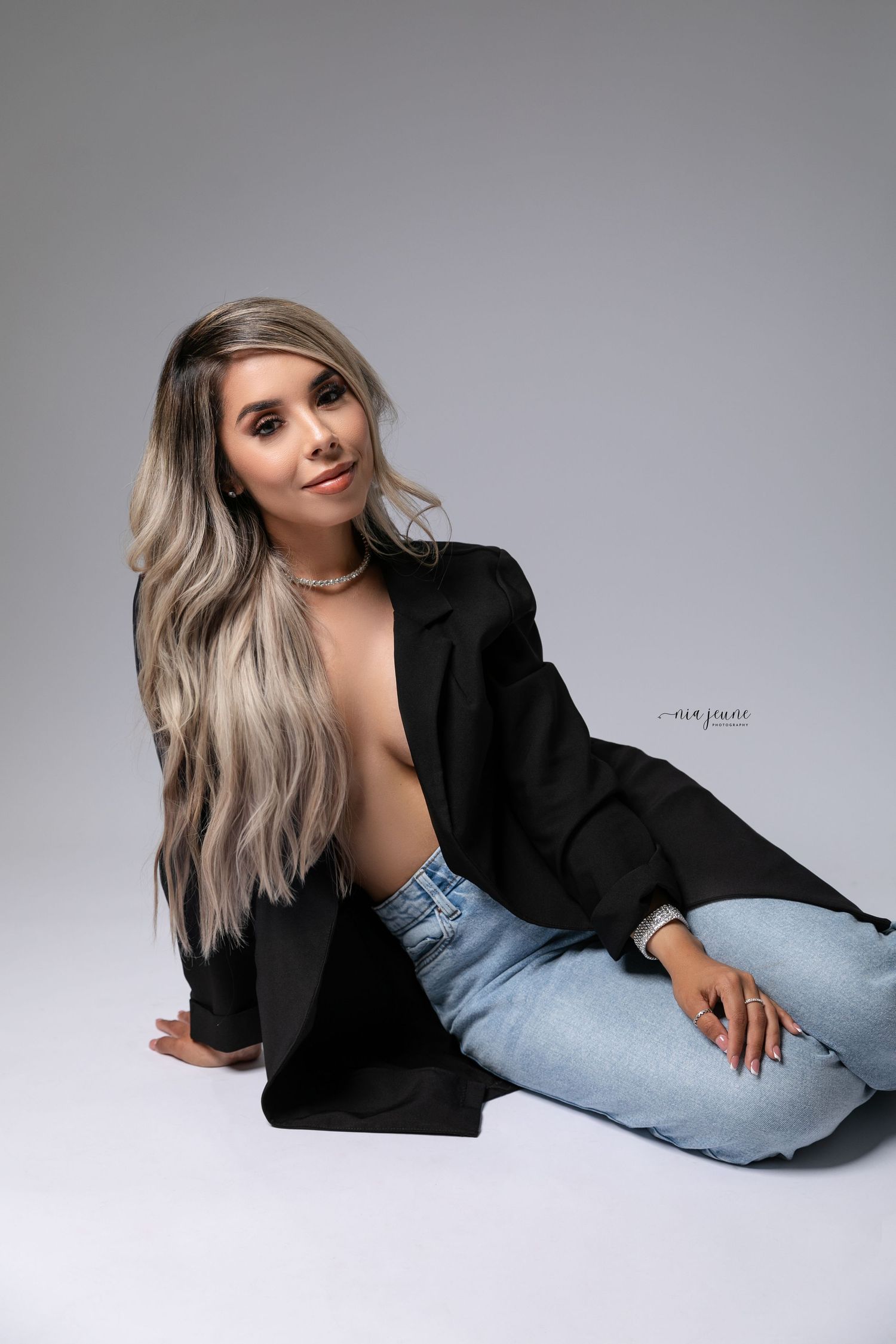 Professional studio portrait of someone in a black blazer and light jeans posing on a grey background.