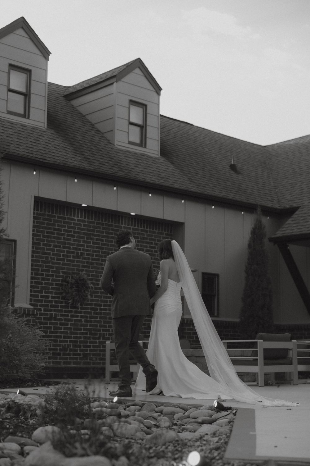 Black and white photo of newlyweds walking toward rustic wedding venue.