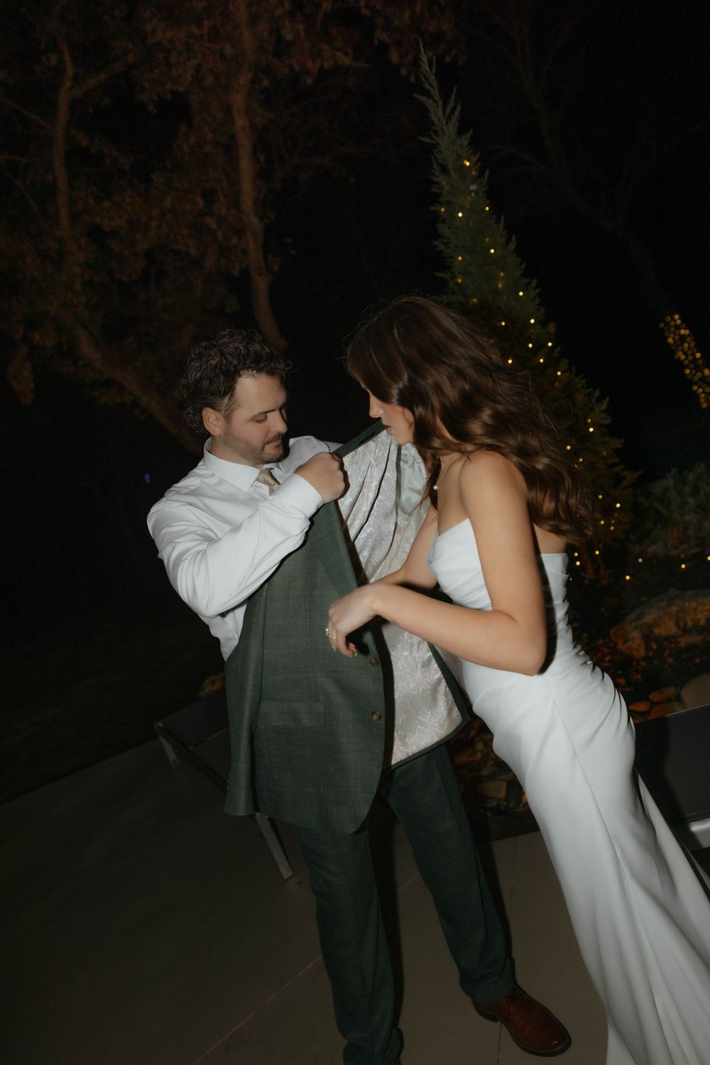 Groom helps bride put on his suit jacket outdoors at night near a lit Christmas tree at wedding.
