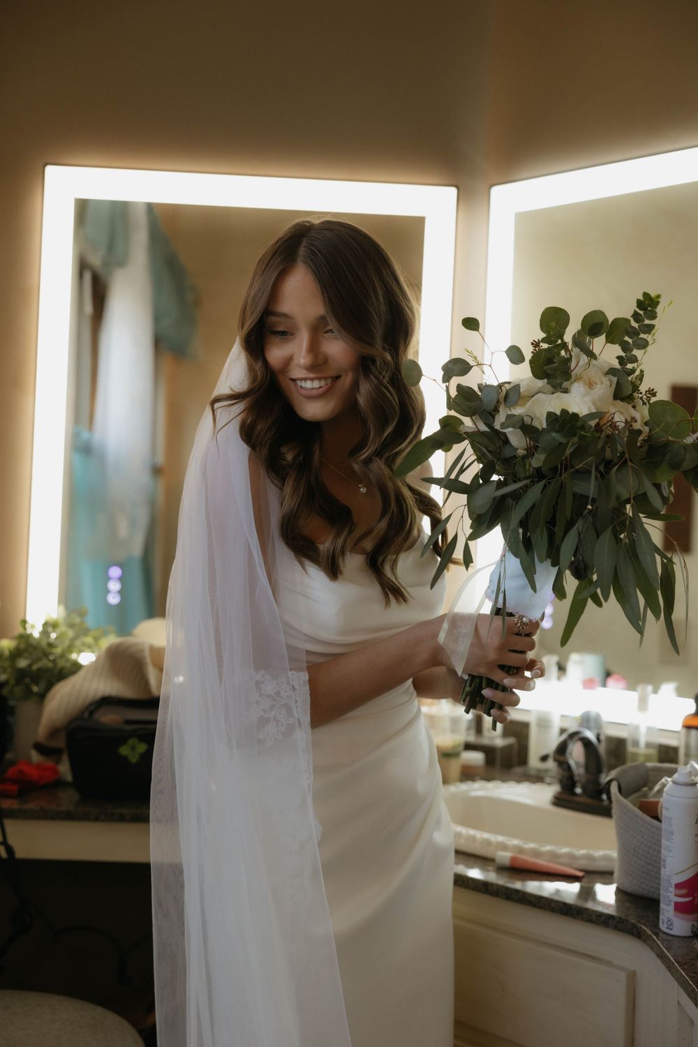 Radiant bride in white gown and veil smiling while holding greenery bouquet in warmly lit bathroom mirror.