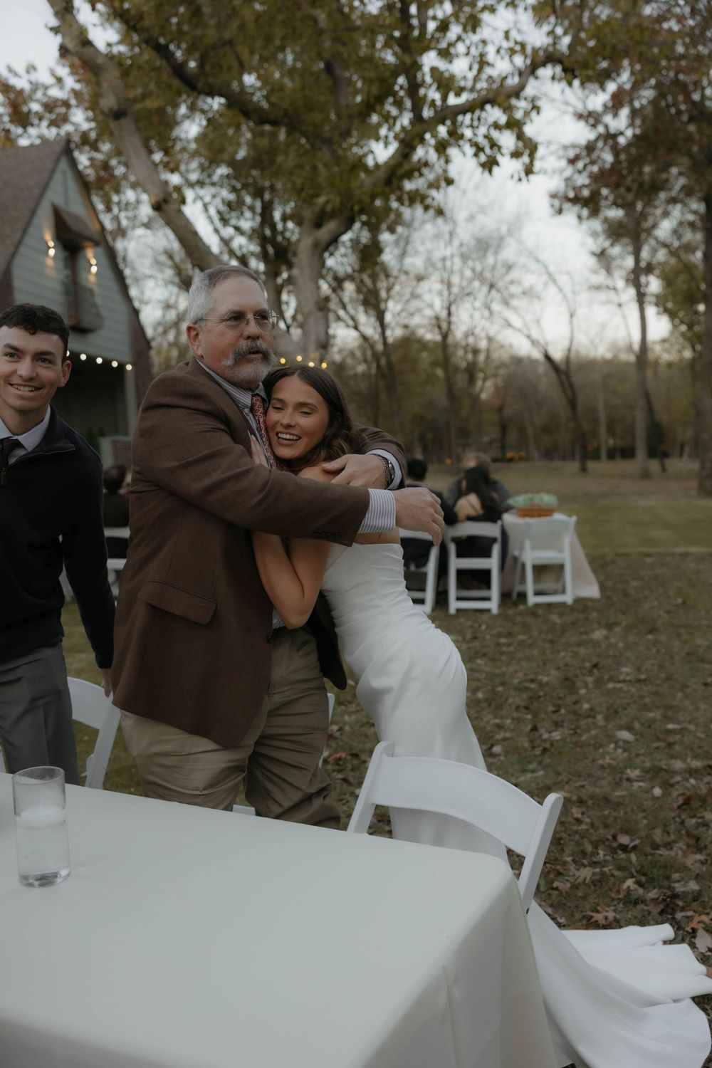 Smiling bride in white gown shares a warm hug with an older man at outdoor wedding reception.