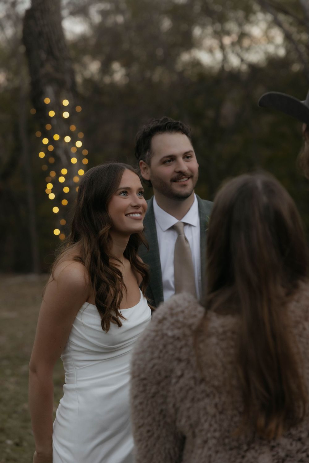 Bride in white dress and groom in gray suit smile while chatting with guests at outdoor wedding.