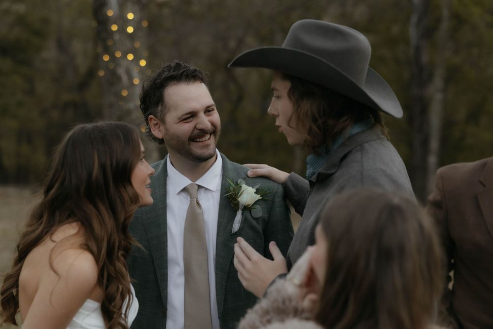 Groom in gray suit laughs with a guest in black cowboy hat at outdoor wedding reception.
