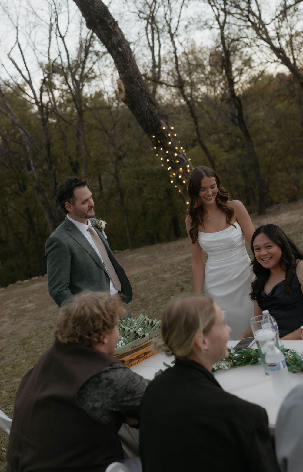 Bride and groom smiling with wedding guests at an outdoor reception table surrounded by trees and string lights.