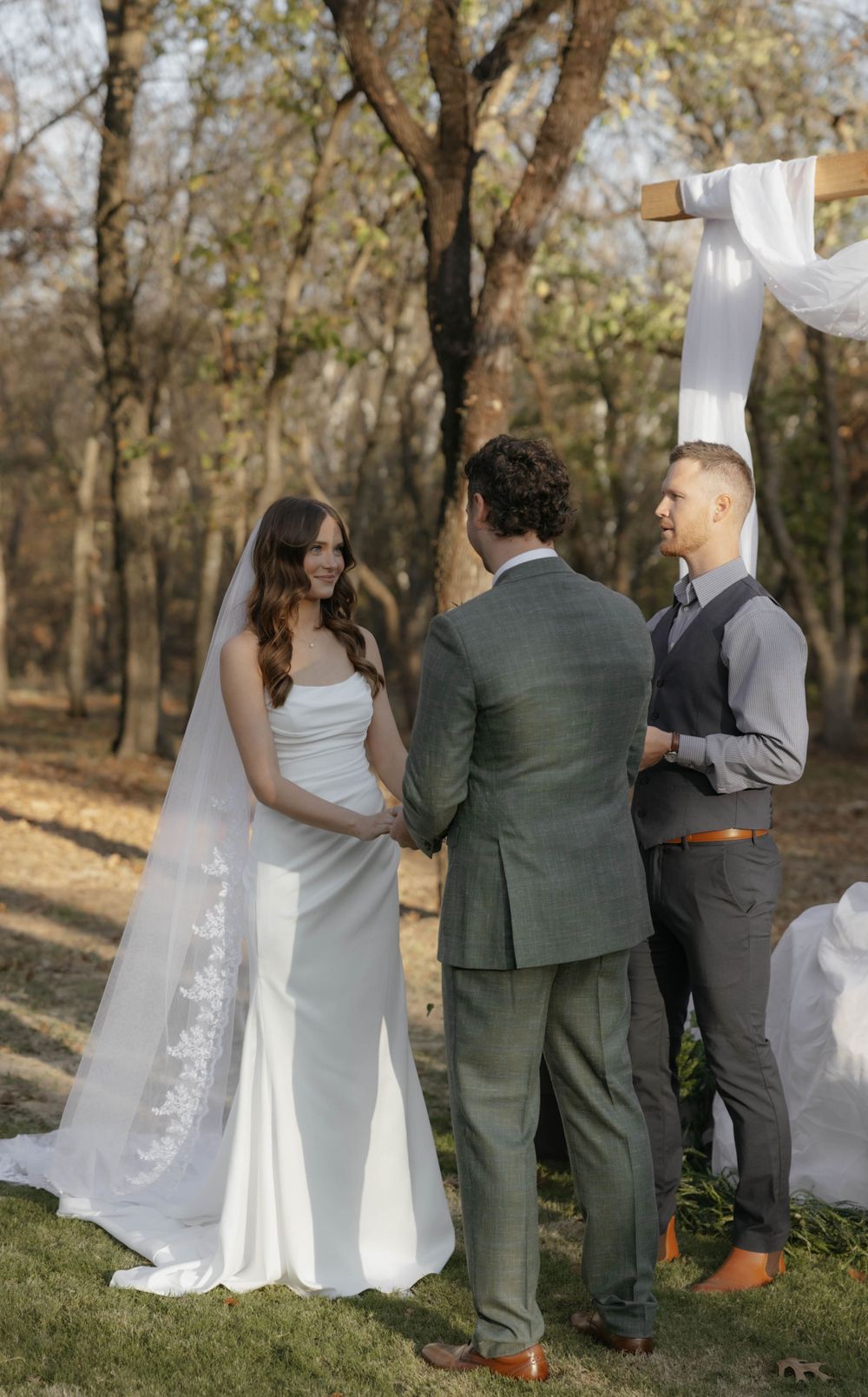 Smiling bride in white gown holds hands with groom at outdoor ceremony.