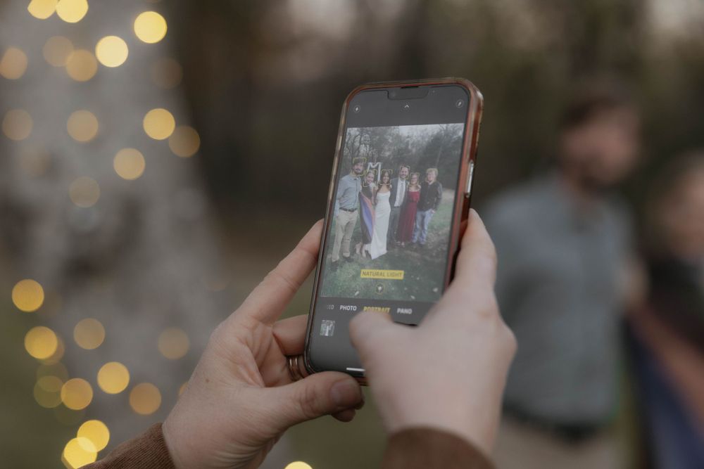 Guest photographs wedding party group photo on smartphone at outdoor evening celebration with bokeh lights.