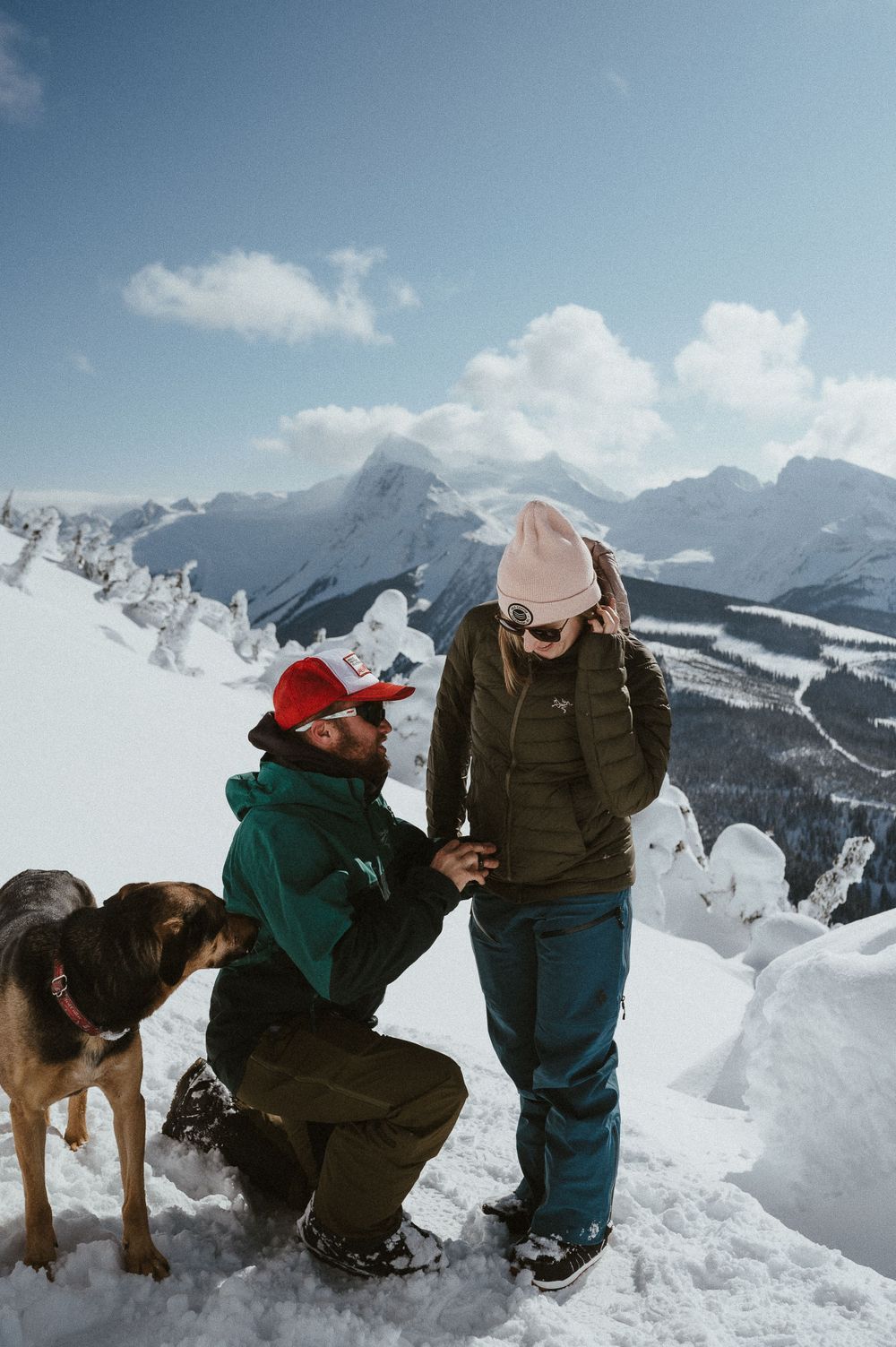A surprise proposal moment unfolds on a snowy mountain peak as a dog watches nearby.