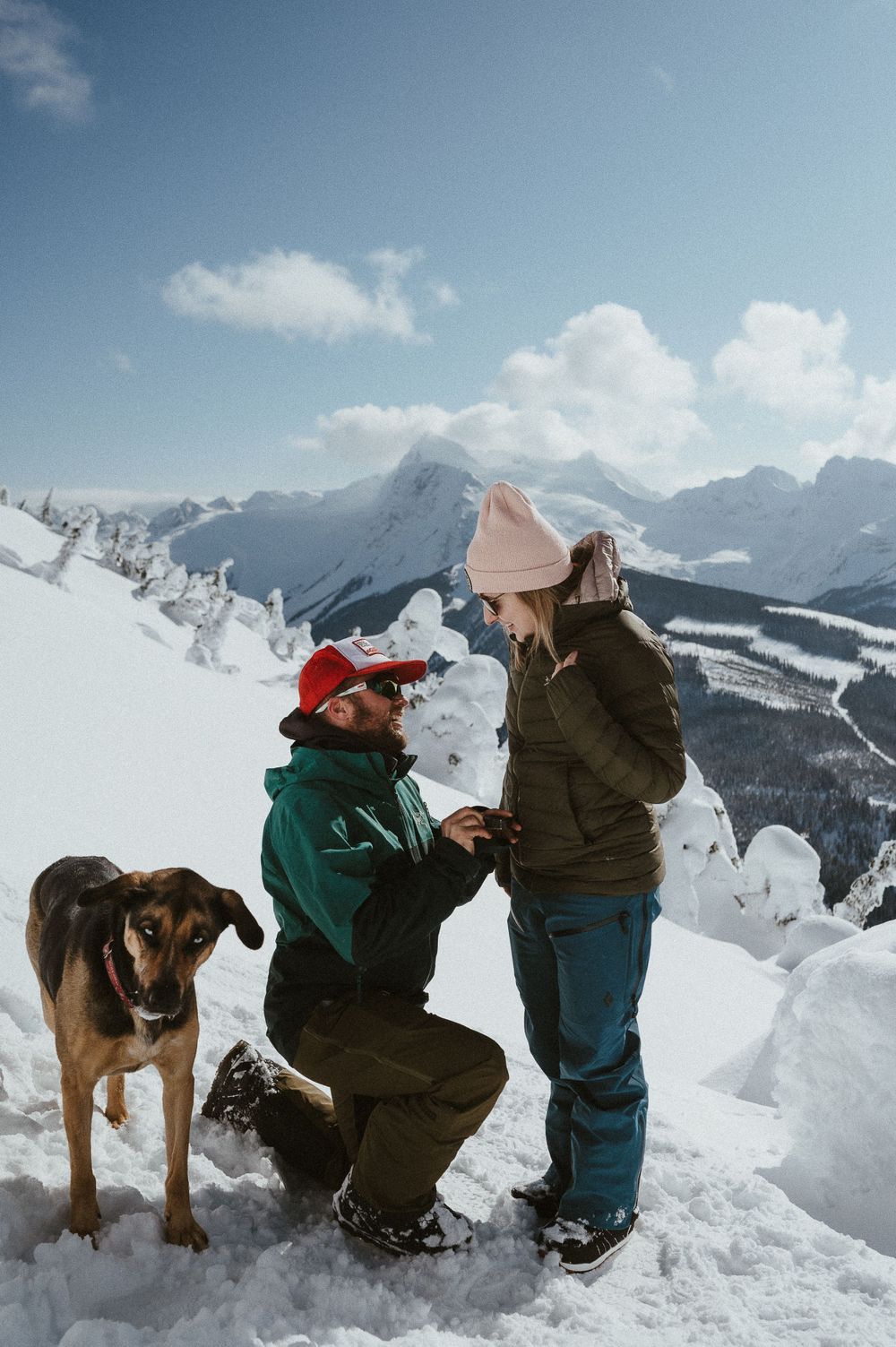 A heartfelt mountain proposal captured against a backdrop of snow-capped peaks with a dog as witness.