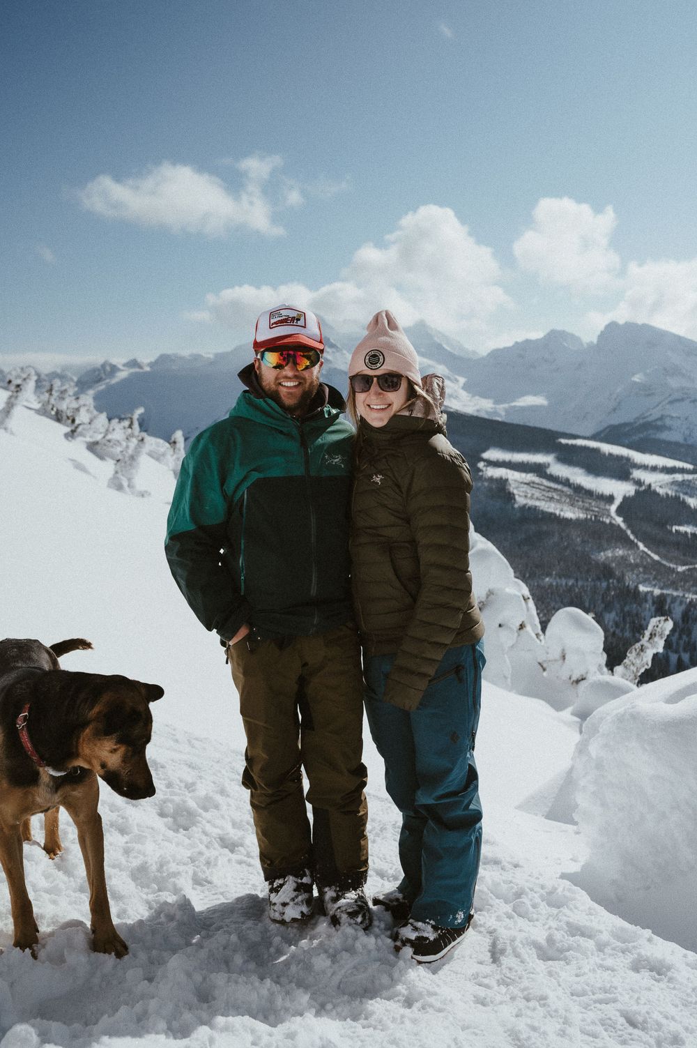 A couple and their dog pose together on a snowy mountain peak with dramatic winter mountain views in the background.