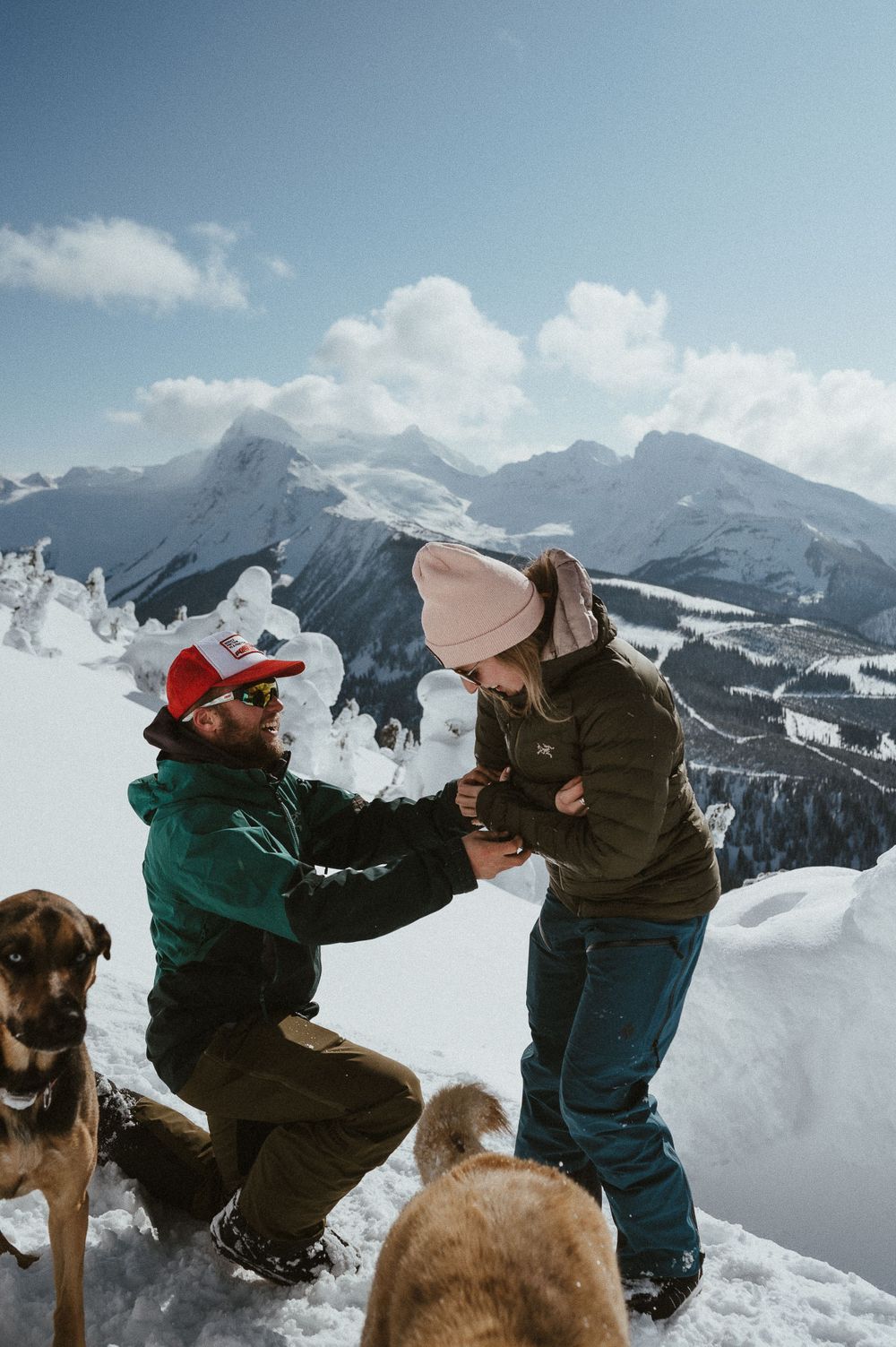 A couple shares a special moment on a snowy mountain peak with their dogs nearby.