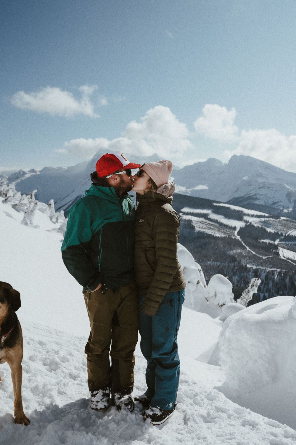 Romantic winter moment shared between two people on a snow-covered mountaintop with their dog nearby.