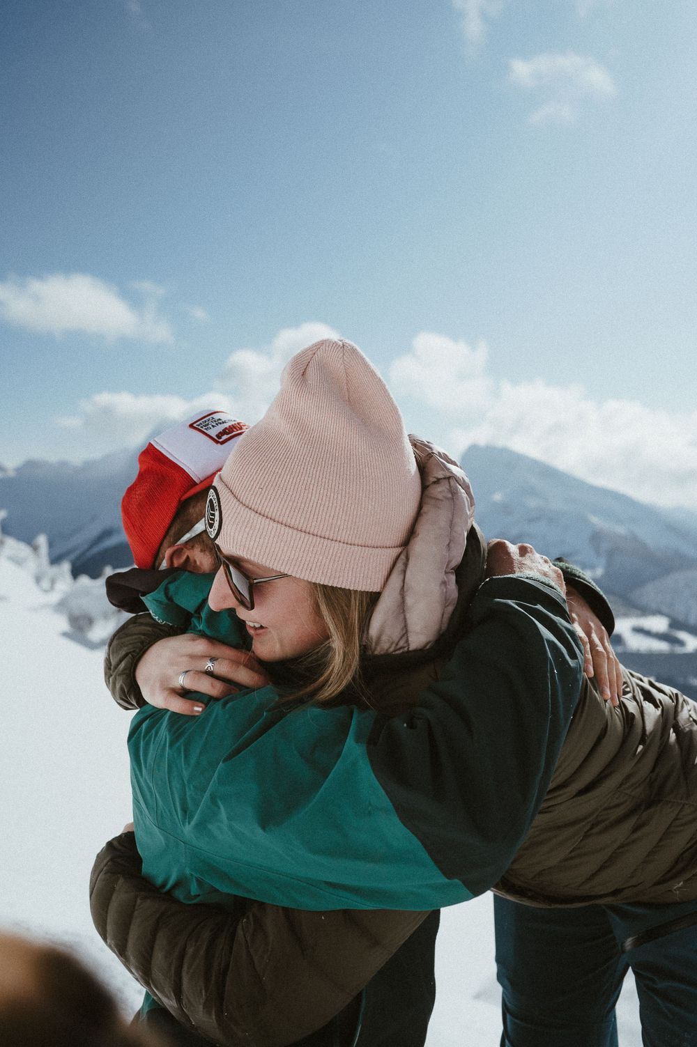 Two people share an emotional hug on a mountain summit with stunning winter views.