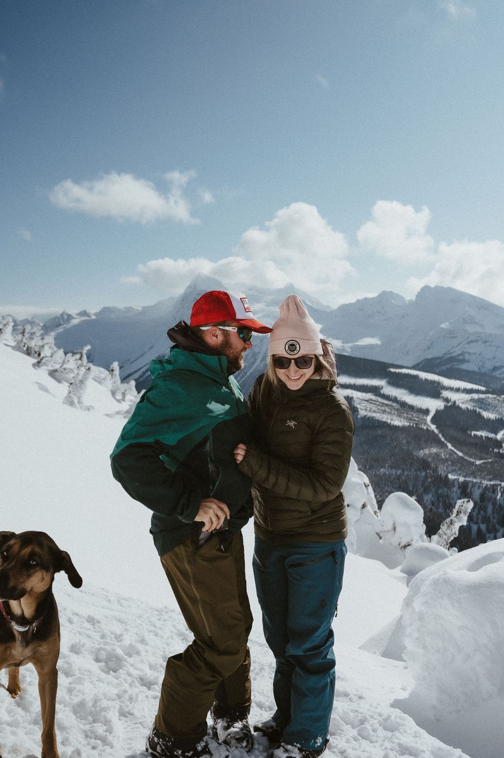 Joyful winter scene with a couple laughing together on a snowy mountain summit while their dog watches.