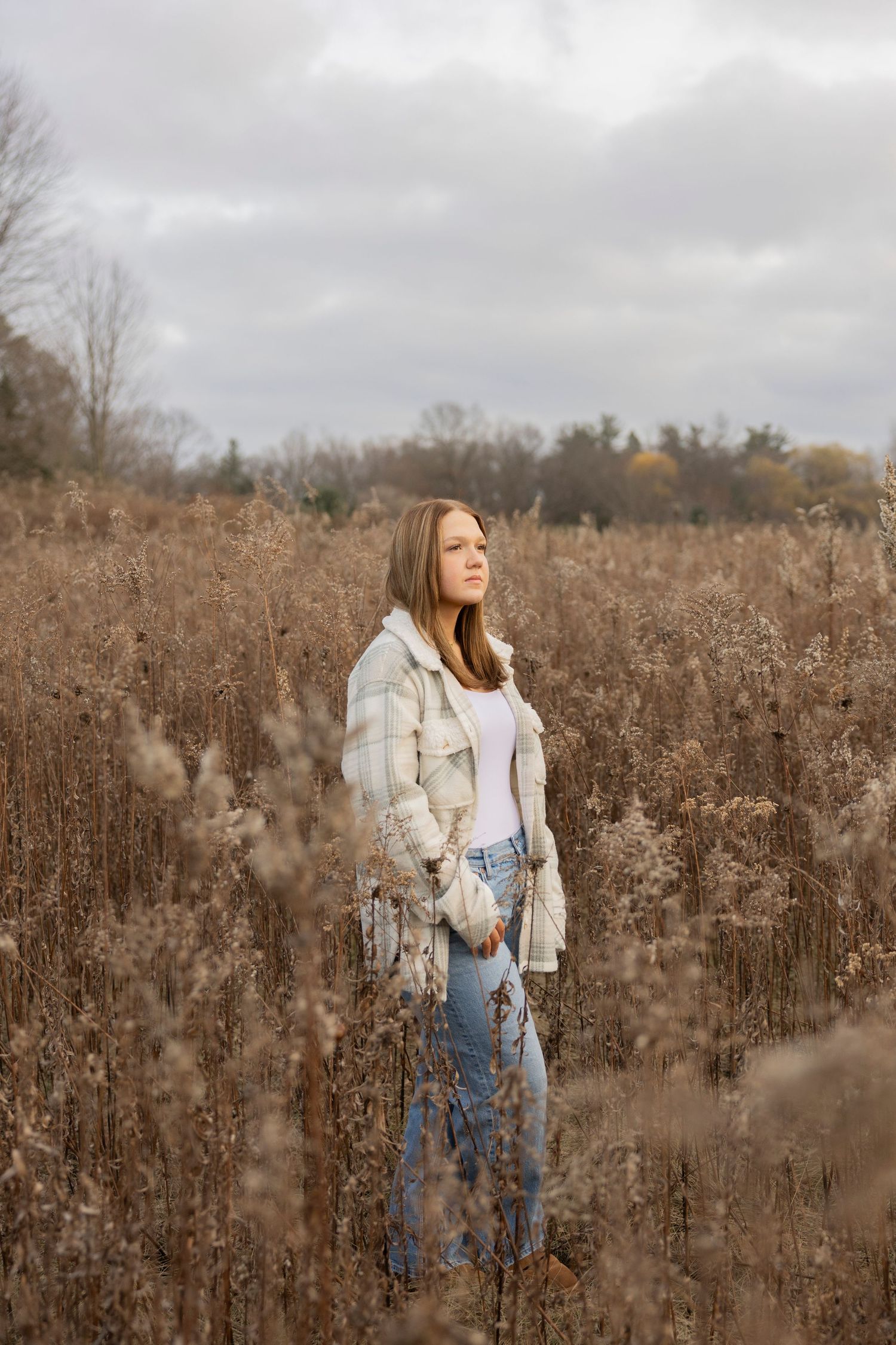 A teenage girl standing in a field of dried autumn grasses wearing a cream sweater and jeans on an overcast day.