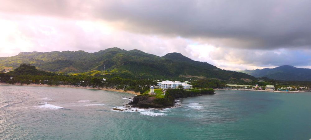 Aerial view of a coastal landscape with mountains and buildings along a tropical shoreline at sunset.