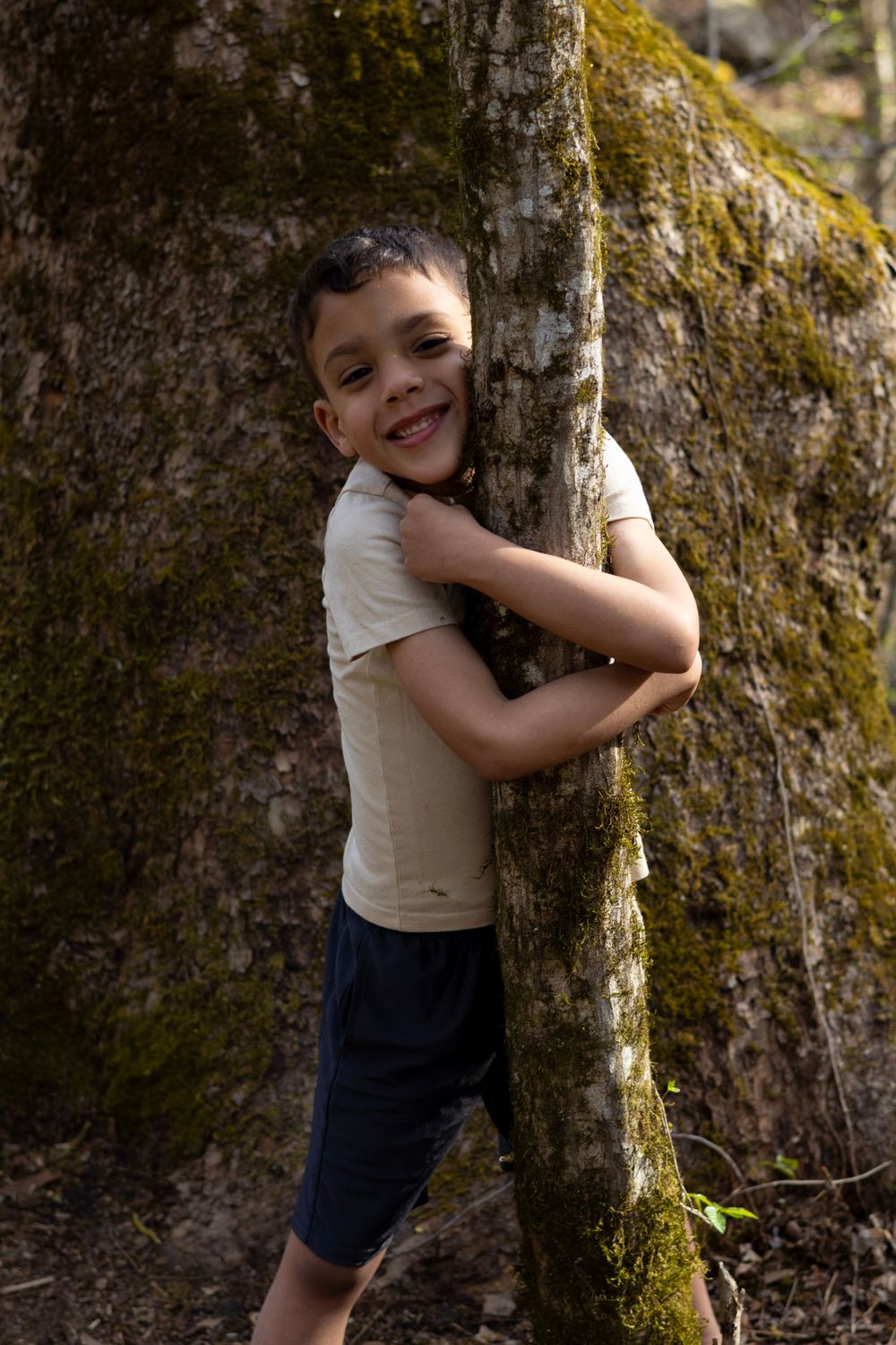 A young explorer hugs a moss-covered tree trunk in a forest setting.
