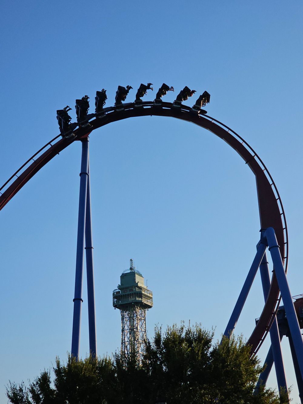 Silhouette of a roller coaster loop against a blue sky with an observation tower in the background.