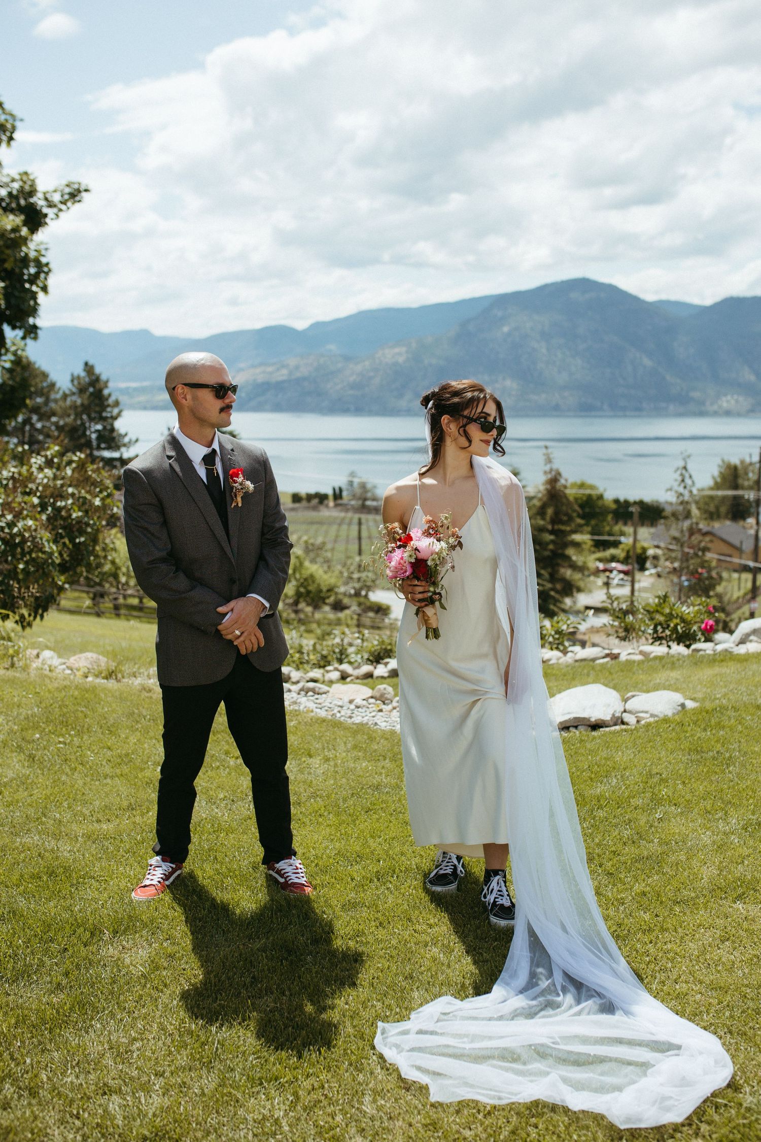 Bride in white midi dress with veil and groom in gray suit stand on green lawn with mountain lake backdrop.