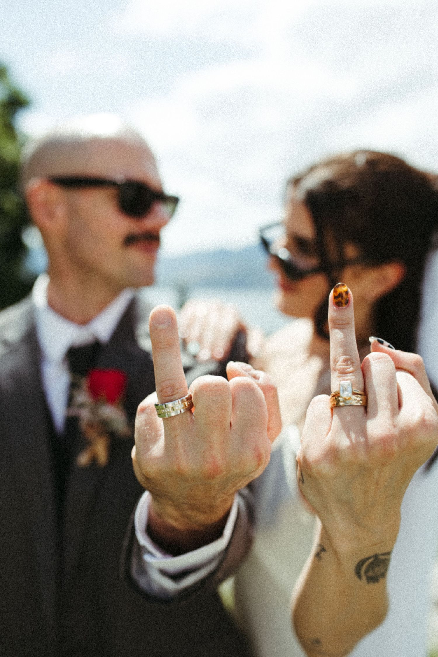 Newlyweds showing off wedding rings on middle fingers, groom in gray suit with rose boutonniere, bride with dark nails.