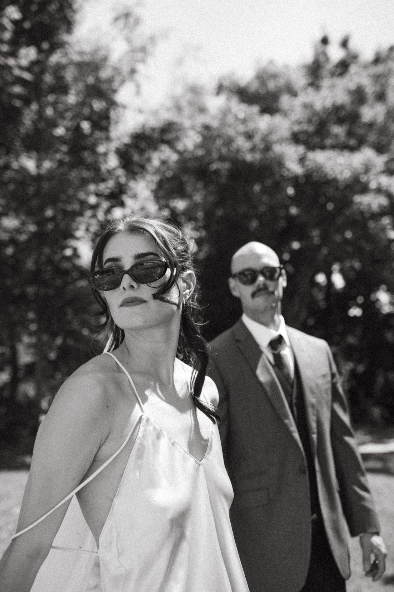 Black and white photo of bride in white slip dress and groom in suit with mustache posing outdoors.