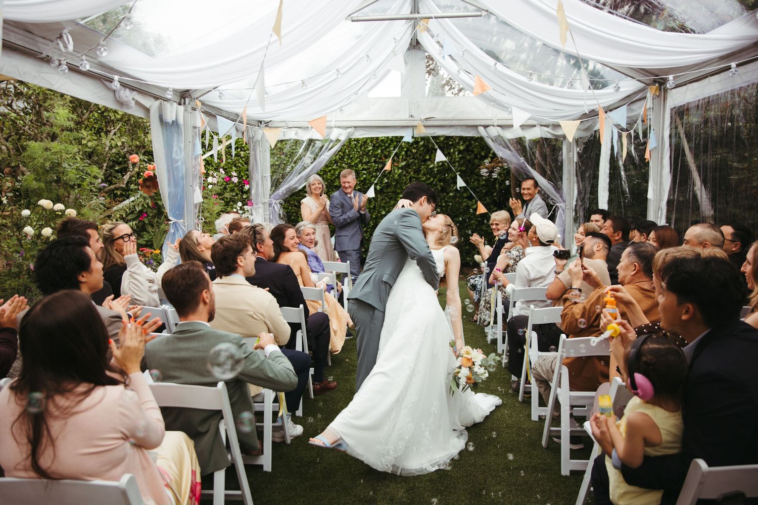 Bride and groom share a kiss under a decorated outdoor tent while wedding guests cheer and toss confetti.