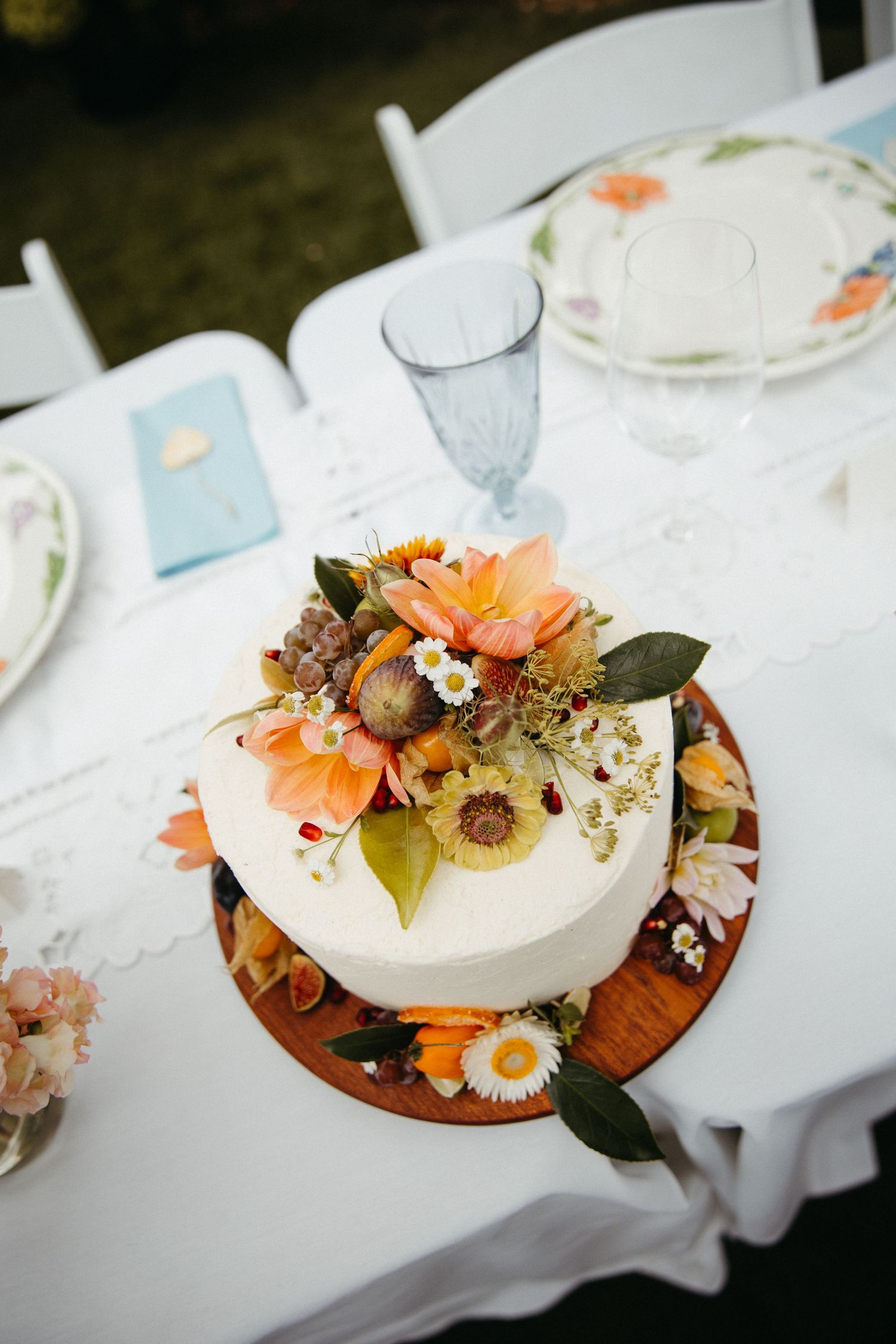 White single-tier wedding cake topped with fresh orange and yellow flowers on a wooden board at an outdoor reception.